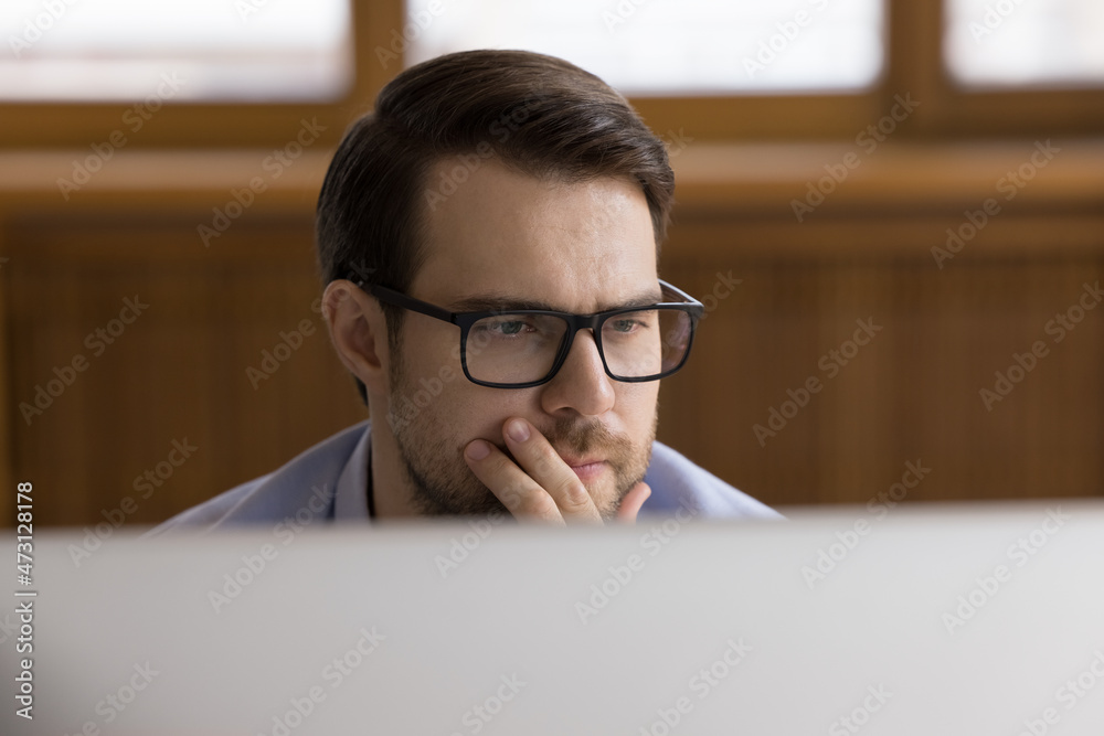 Poster serious male employee in glasses working at office workplace, sitting at desktop monitor, reading, w