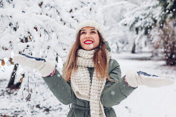 Happy young woman playing with snow in snowy winter park wearing warm knitted clothes and having...