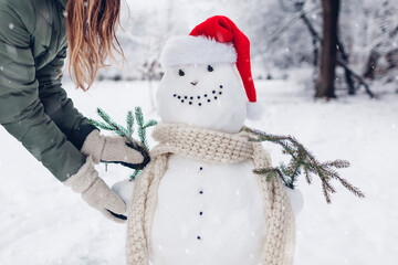 Girl makes snowman outdoors in snowy winter park dressing it in Santa hat, scarf and attaching fir...