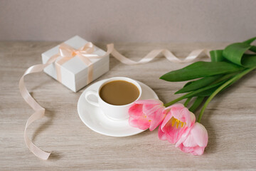 Coffee with milk in a white porcelain cup and saucer, a bouquet of spring pink tulips and a gift in a white box with a satin ribbon. Mother's Day, Valentine's Day, Easter. The concept of spring