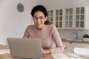 Smiling woman in glasses using laptop, sitting in kitchen at home, happy young female looking at computer screen, watching webinar or movie, positive businesswoman freelancer working on project