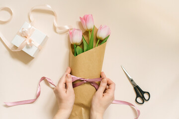 Flat female hands tie a satin ribbon bow on a simple bouquet of fresh pink tulip flowers in craft paper on a beige table.