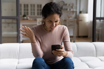 Confused angry woman looking at smartphone screen, sitting on couch at home, dissatisfied shocked...