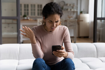 Obraz premium Confused angry woman looking at smartphone screen, sitting on couch at home, dissatisfied shocked young female having problem with discharged or broken device, reading spam message or bad news