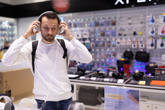 Photo of an adult handsome thinking brunet male person with stubble in a white sweatshirt chooses earphones in an electronic store in a shopping mall. Shop and music concept