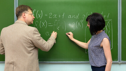 An adult teacher at the blackboard explains the solution of a math problem to his student.