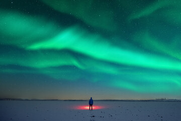 Tourist with red flashlight on snowy field against the backdrop of incredible starry sky with Aurora borealis. Amazing night landscape. Northern lights in winter field - obrazy, fototapety, plakaty
