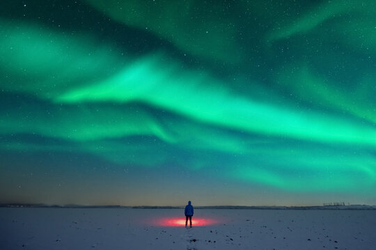 Tourist with red flashlight on snowy field against the backdrop of incredible starry sky with Aurora borealis. Amazing night landscape. Northern lights in winter field