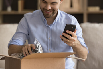 Happy consumer man unpacking parcel with Christmas gifts, producing wrap with sweater from...