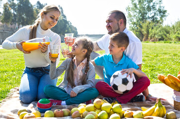 mother pours orange juice into glasses to husband, daughter and son at picnic in park