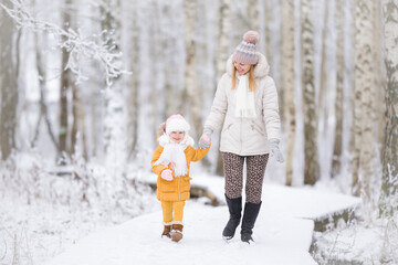 Happy little daughter and young adult mother walking on white snow covered trail at birch tree...