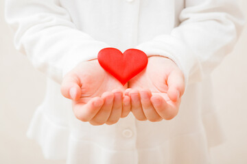 Baby girl hands holding and showing bright red heart shape. Toddler in white clothes. Closeup. Front view. Love concept.