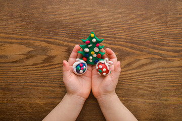 Green Christmas tree with colorful balls and gift boxes created from modeling clay in baby girl palms on brown wooden table background. Closeup. Child dreaming about presents. Top down view.