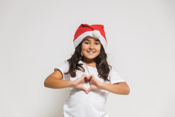 Smiling child girl in christmas santa hat showing heart gesture, congratulating with xmas holiday. Love, care and trust