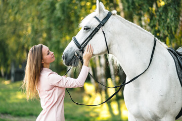 Smiling young female caring about horse outdoors. Happy girl stroking white thoroughbred equine. Pet therapy concept