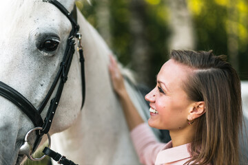 Smiling young female caring about horse outdoors. Happy girl stroking white thoroughbred equine. Pet therapy concept