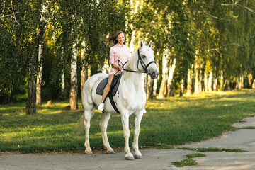Happy young girl practicing horseback riding on thoroughbred white horse outdoors. Equestrianism school advertising