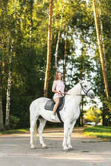 Happy young girl practicing horseback riding on thoroughbred white horse outdoors. Equestrianism school advertising