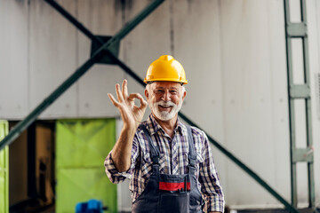 A happy senior blue-collar employee with a protective helmet is standing in the factory and showing...