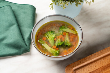 Small portion of kids vegetarian soup with broccoli and carrot cubes, light kitchen table with napkin and cutting board aside