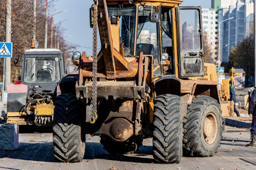A heavy front loader transports earth in a bucket and grades the road. Road repair in the center of a modern city in autumn. Heavy construction machinery for moving soil.
