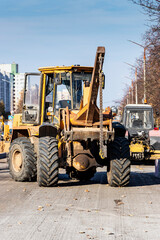 A heavy front loader transports earth in a bucket and grades the road. Road repair in the center of a modern city in autumn. Heavy construction machinery for moving soil.