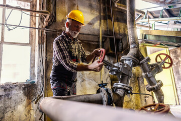 An old factory worker with a helmet on his head is screwing a valve in the factory. Factory worker...