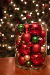 Holiday table center piece of a glass jar filled with Christmas ornaments and a lit tree in the background.