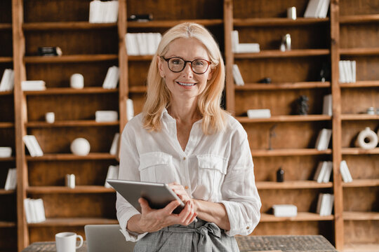 Smart mature middle-aged businesswoman freelancer tutor teacher boss ceo holding digital tablet looking at the camera while working remotely in home office