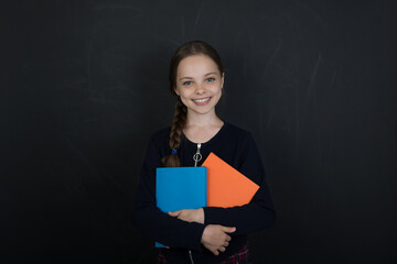 Happy cute child girl  holding books. Smilling schoolgirl   front of black chalkboard.