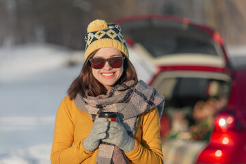 Woman with coffee in hands is sitting in car trunk and has winter picnic