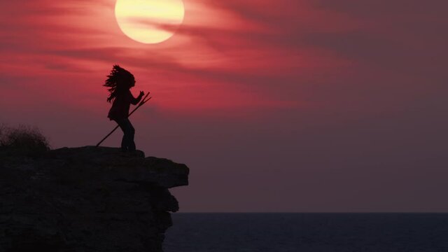 Silhouette of Red Indian chief standing on edge of mountain near ocean or sea calls on people against backdrop of amazing sunset sky with large sun.