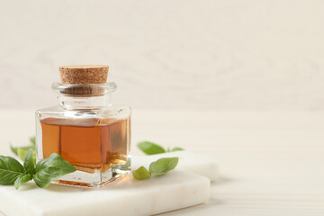 Bottle of essential basil oil and fresh leaves on white wooden table, space for text