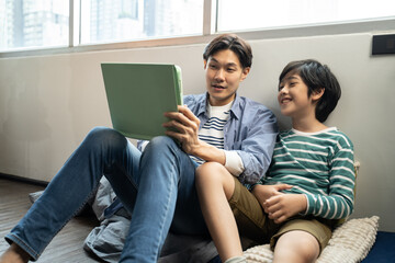 Cheerful asian Father And Son sitting on floor together and Watching Funny Videos On Digital Tablet.Asian homeschool boy sitting with father, learn online by using tablet concept.