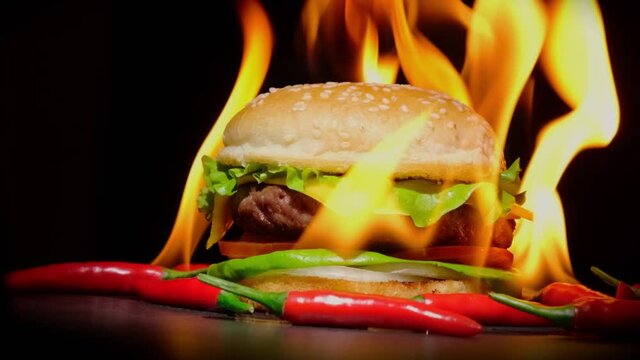 Juicy spicy beef burger with red hot chili peppers on fire rotating on table on black background close-up