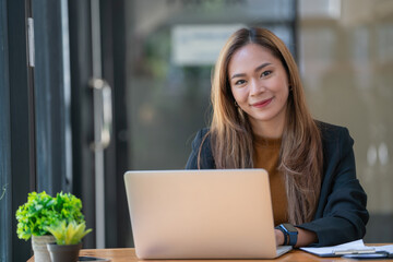 Portrait of a young businesswoman using a laptop, smiling and looking at camera while sitting at office desk in modern office.