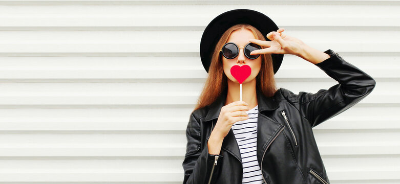 Fashionable portrait of stylish young woman with red heart shaped lollipop blowing her lips sending sweet air kiss wearing a black round hat, leather jacket on white background