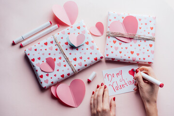 Top view of female hands on pink background with gifts writing valentine card BE MY VALENTINE