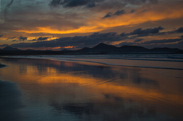 Obraz premium Sunset at Famara Beach. Mountains in the background and orange clouds reflecting in the wet sand. Lanzarote, Spain.