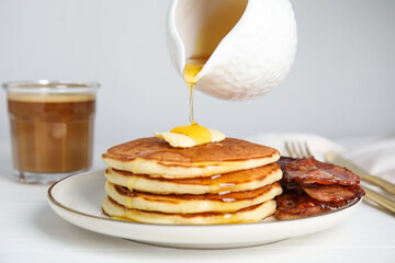 Pouring maple syrup onto delicious pancakes with butter and fried bacon at white wooden table,...