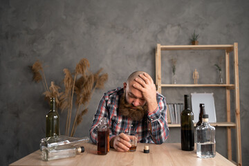 adult bearded man sitting at the table drinking a bottle of alcohol at home sad alone, alcoholism,...