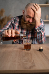 Depression and alcoholism in men. An adult bearded guy himself sits at a table with bottles of alcohol. He holds brandy in his hands and pours it into a glass.