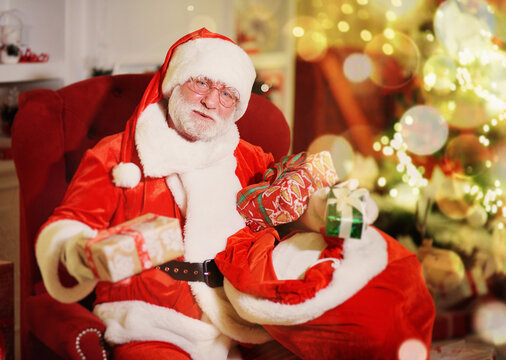 Santa Claus is smiling sitting on a red armchair and holding a bag of gifts against the background of a Christmas tree