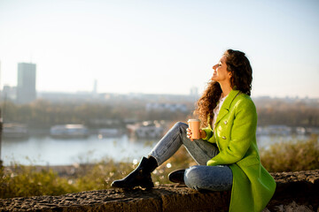 Pretty young woman enjoying autumn sun while sitting by the river and drinking takeaway coffee