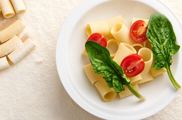 Flat lay of a vegetarian uncooked traditional Italian whole wheat pasta with halves of tomato and green spinach leaves on a floured background with scattered raw tortiglioni