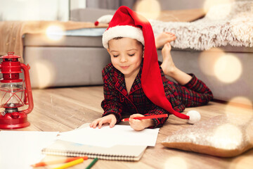 Little boy child in a santa hat writes a letter to santa claus lying on the floor.