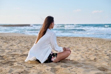Young relaxed woman sitting in lotus position on beach, morning sea nature