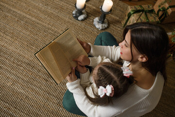 A little daughter and mother in white sweaters are reading a book, near the Christmas tree. top view