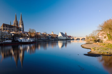 Fototapeta na wymiar Donauufer in Regensburg mit Dom, Spiegelungen und steinerner Brücke bei blauem Himmel, im Abendlicht im Winter