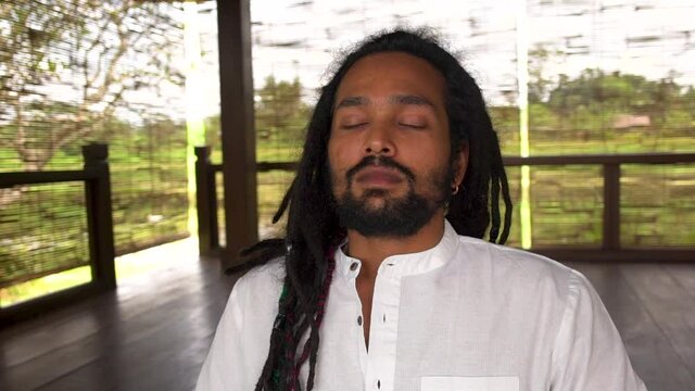 Close-up of indian man with dreadlocks do meditation practice, young man wearing white shirt sitting at yoga place with closed eyes. Dollyshot.